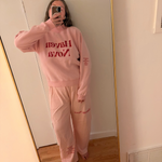 A person taking a mirror selfie in a matching light pink lounge set with red text reading 'Haven Nova' on the sweatshirt and pants, standing in a room with a wood floor and white wall. 12
